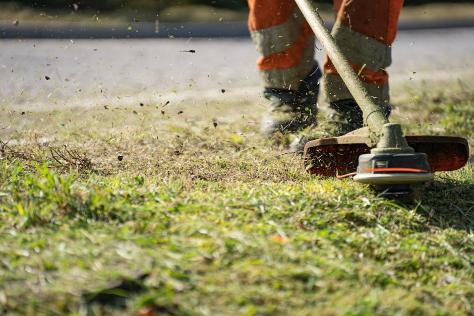 Close-up of a gardener's legs and feet wearing orange work trousers, operating a grass trimmer in a well-maintained garden. The garden features a green lawn with patches of freshly cut grass scattered