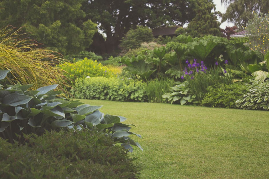 Close-up of a gardener's legs and feet wearing orange work trousers, operating a grass trimmer in a well-maintained garden. The garden features a green lawn with patches of freshly cut grass scattered